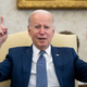 President Joe Biden sits in a chair with a blue suit and tie, raising his arms and pointing upwards while in the Oval Office.